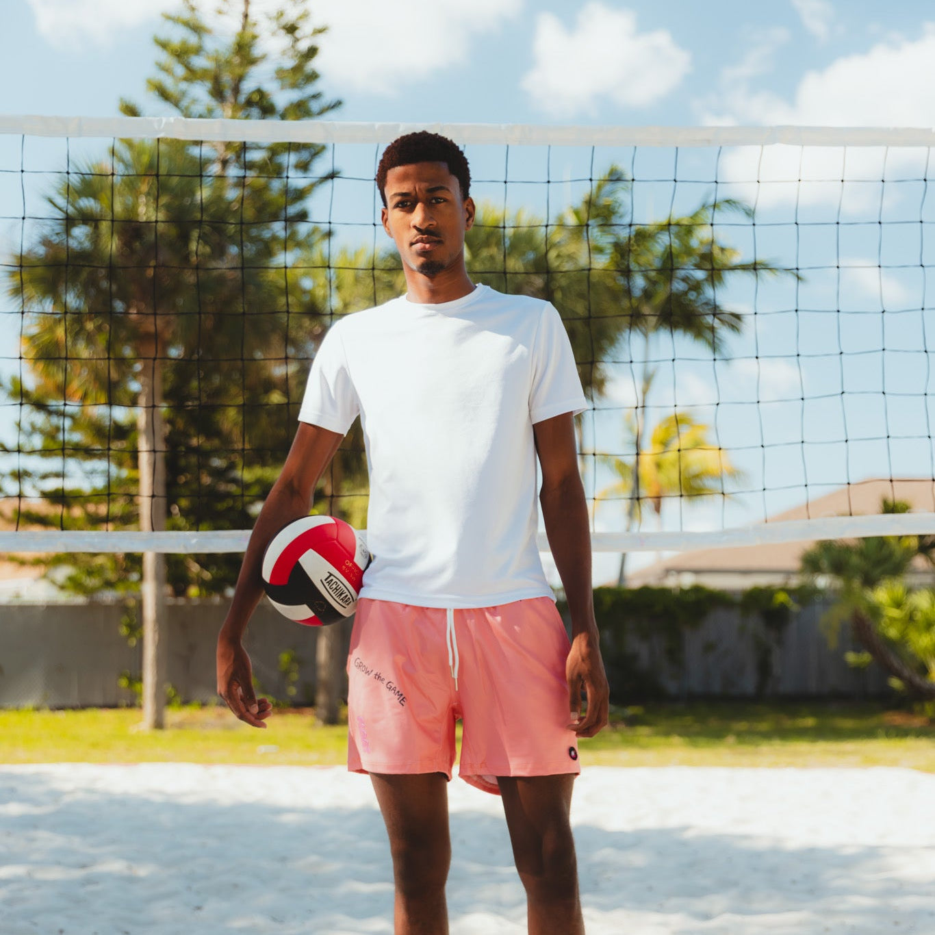 Man holding a volleyball on a sandy court with a net and trees in the background