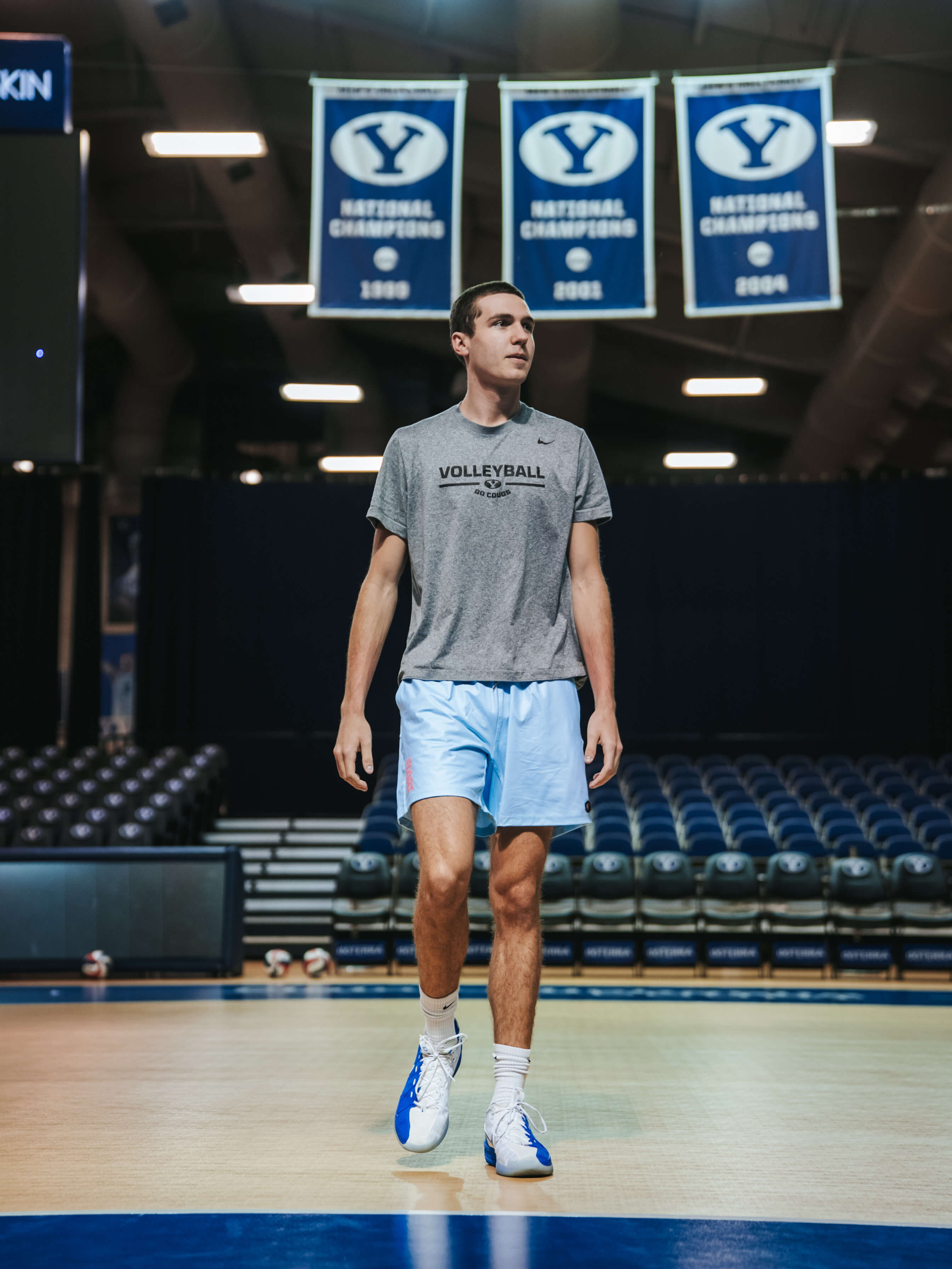 Man in gray 'Volleyball' t-shirt and light blue shorts standing on a volleyball court with championship banners in the background.