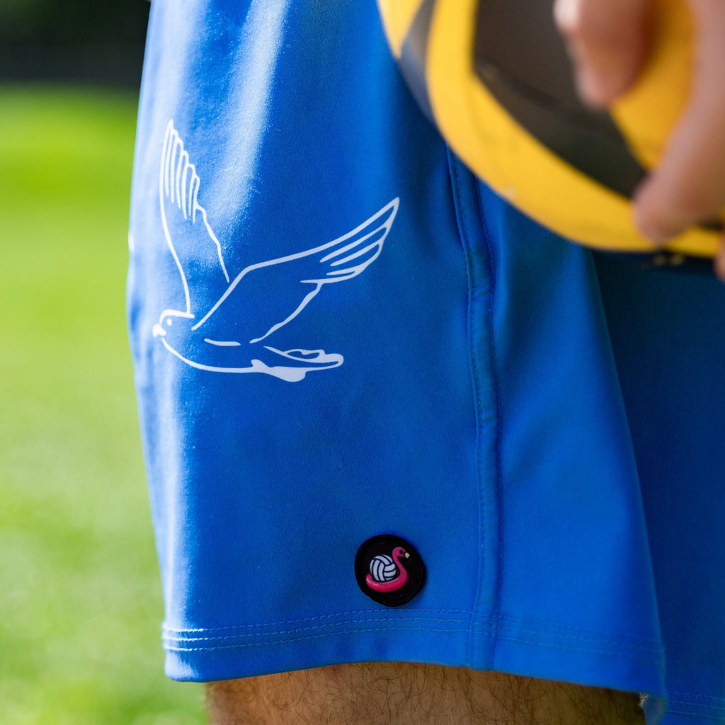 Close-up of a person wearing bright blue athletic shorts with a white bird outline logo and a small circular black logo near the hem, holding a yellow and black ball outdoors on a grassy field.