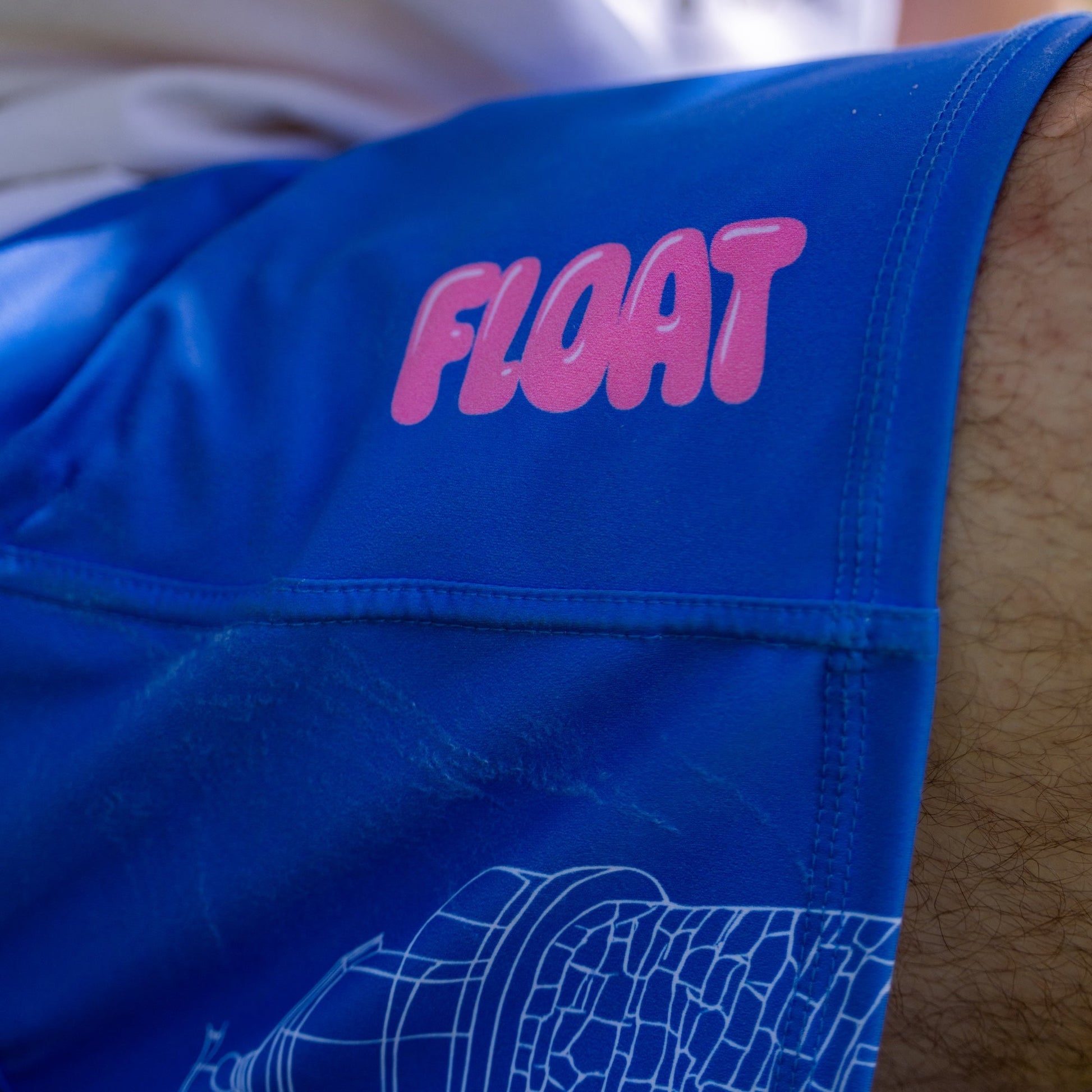 Close-up of a person wearing bright blue swim shorts with a pink 'FLOAT' logo on the hip and a white line-art graphic near the hem.