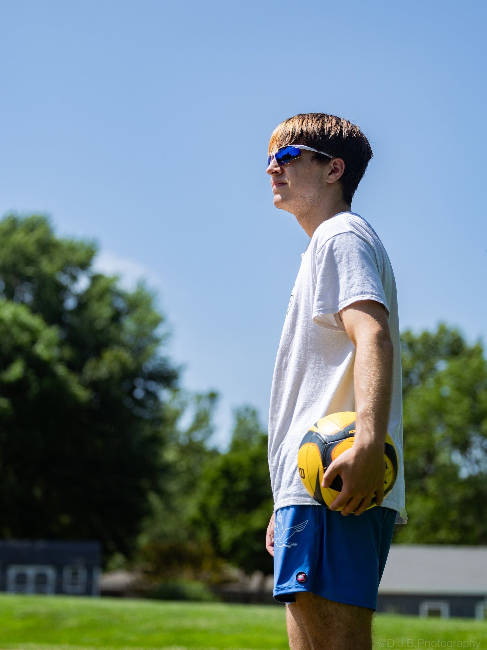 A young man in a white t-shirt, blue shorts, and blue sunglasses stands on a grassy field holding a yellow soccer ball under a clear blue sky.