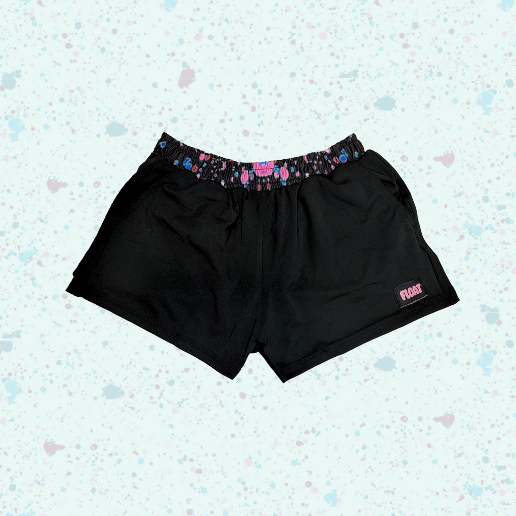 Black athletic shorts with a colorful patterned elastic waistband and a small pink logo on the lower left leg, displayed against a light blue speckled background.