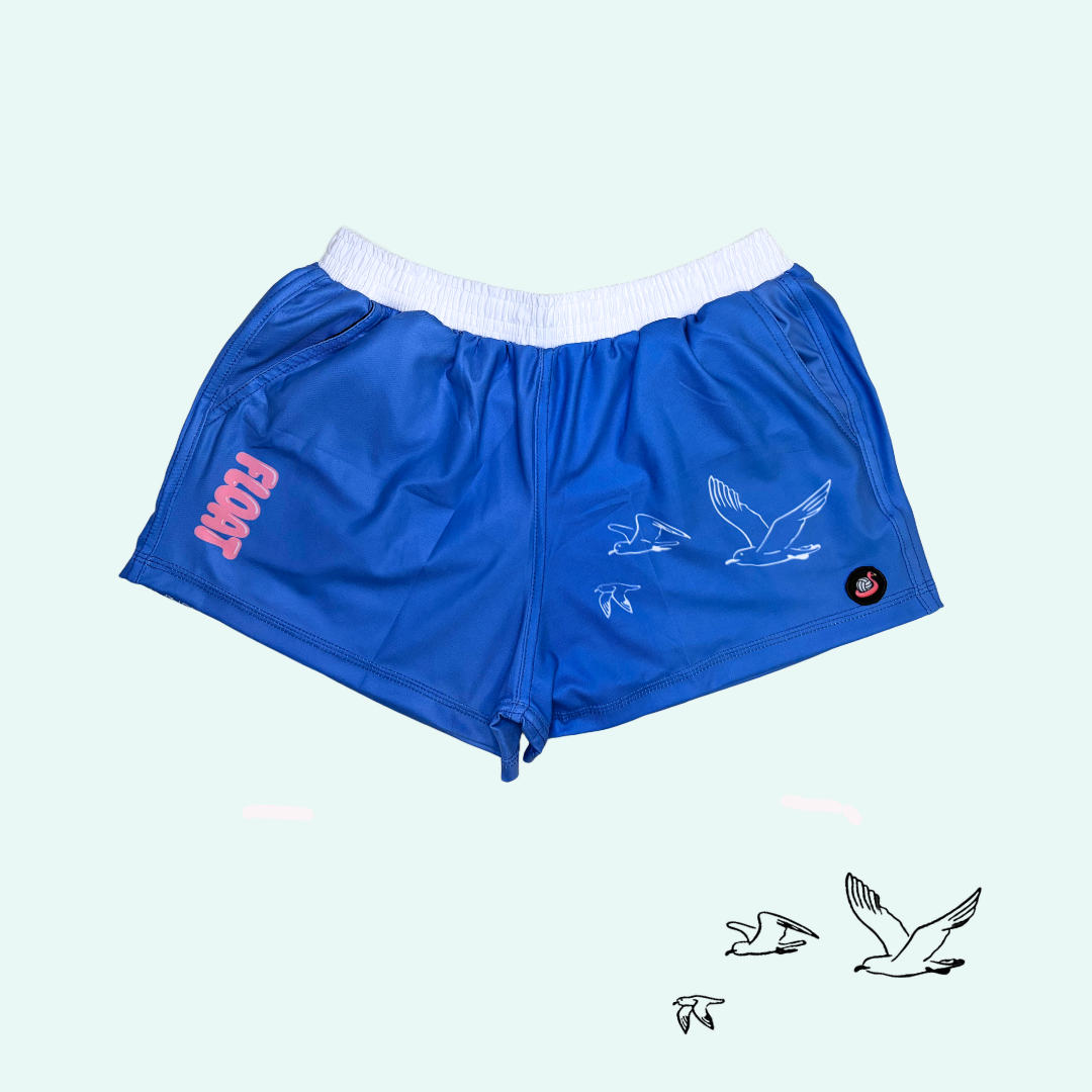 Blue athletic shorts with a white waistband, featuring a pink 'FLOAT' logo on the left leg, a white bird graphic on the right leg, and a small black circular patch on the lower right side, shown against a pale mint green background.