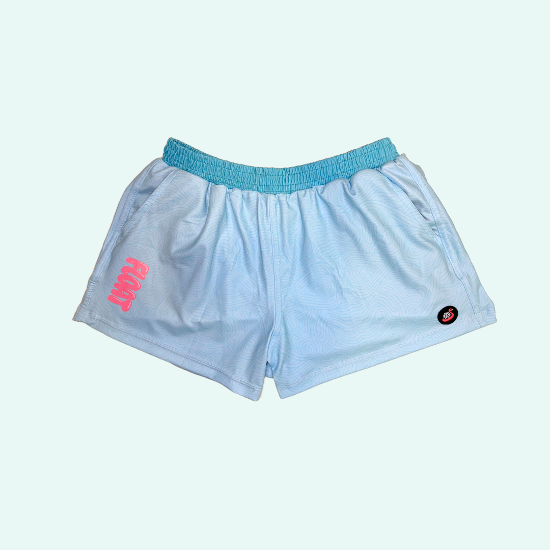 Light blue athletic shorts with a turquoise elastic waistband, featuring a pink logo on the left leg and a small black circular logo on the right leg, displayed against a pale mint green background.