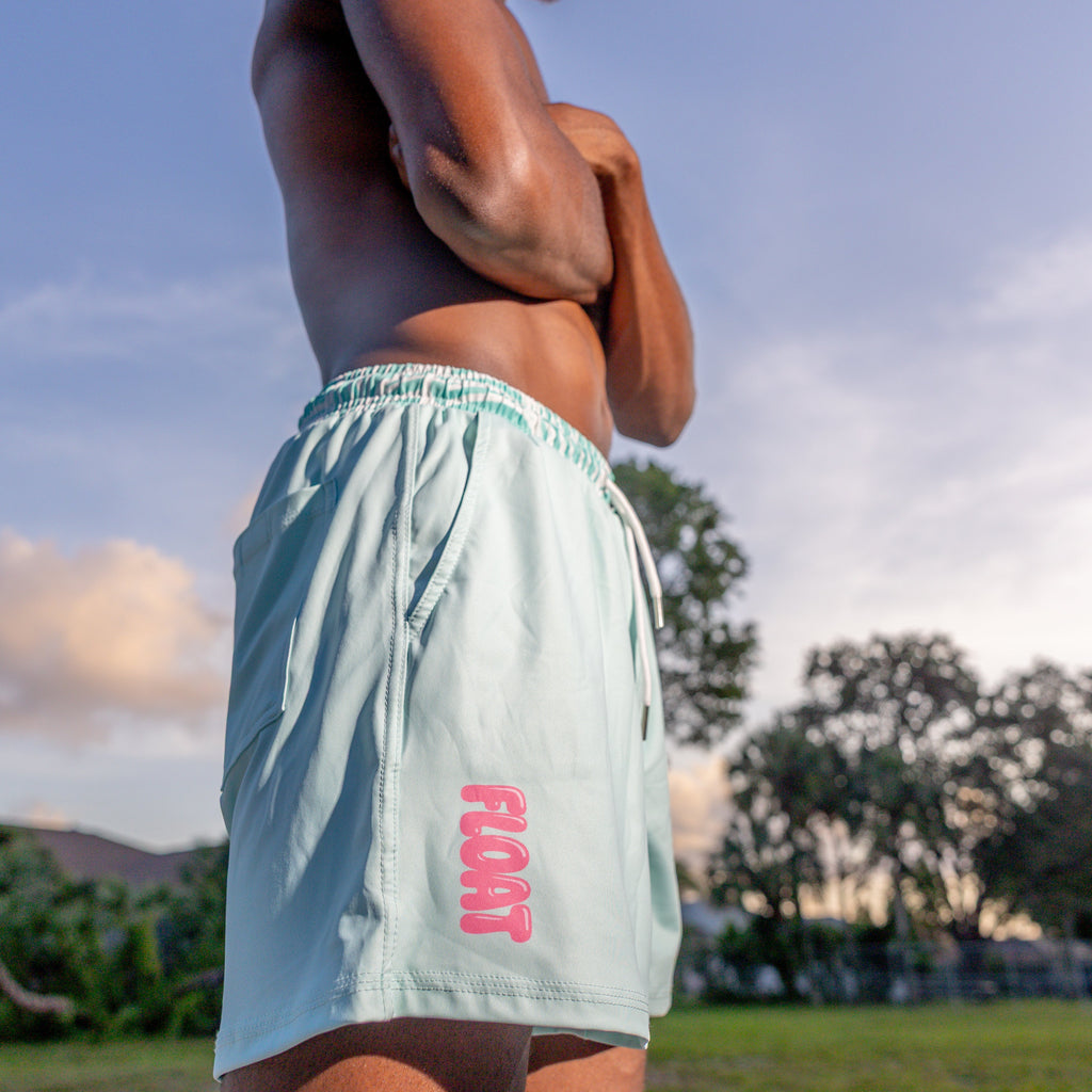 Man wearing light blue shorts with 'FLOAT' logo standing on a beach with trees and sky in the background