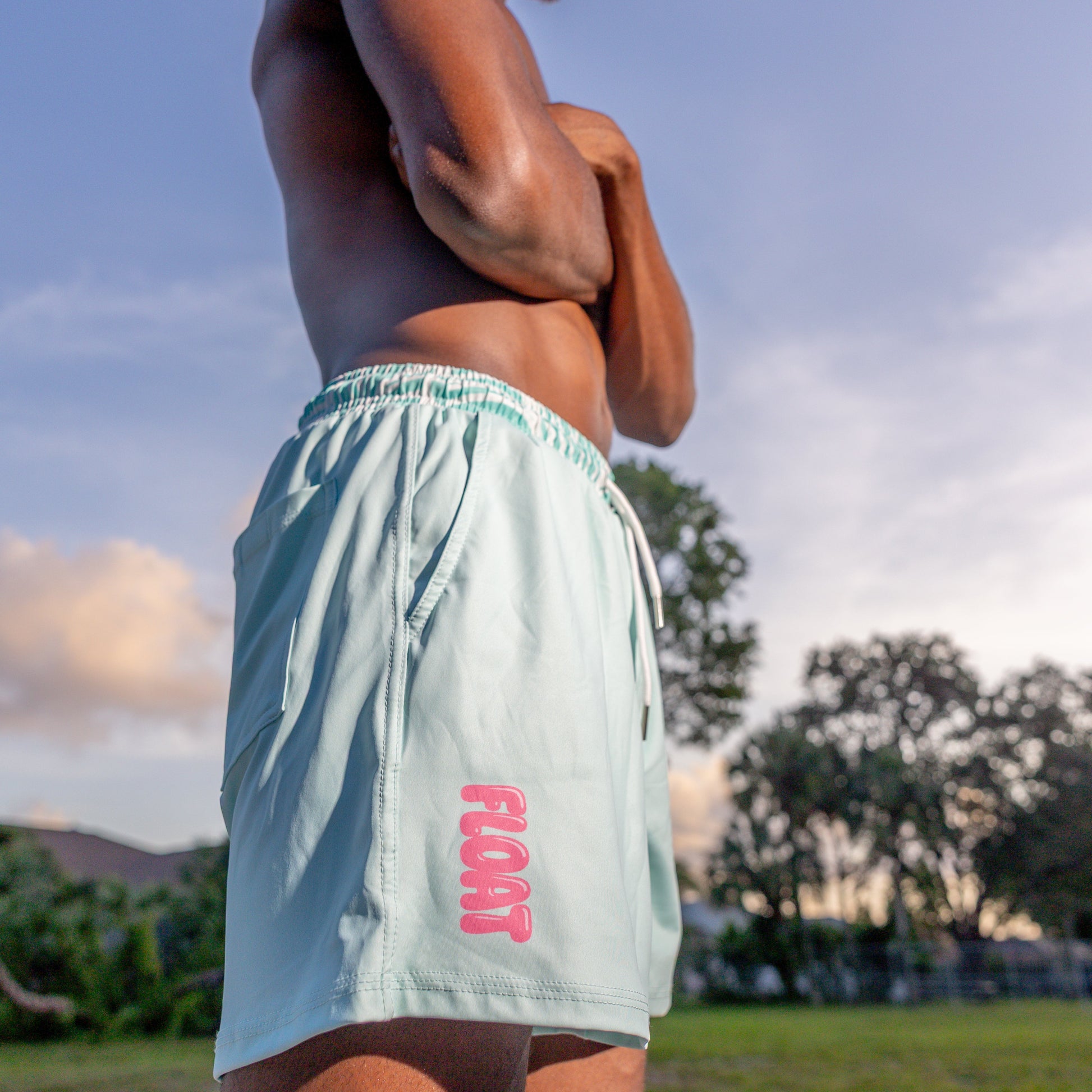 Man wearing light blue shorts with 'FLOAT' logo standing on a beach with trees and sky in the background