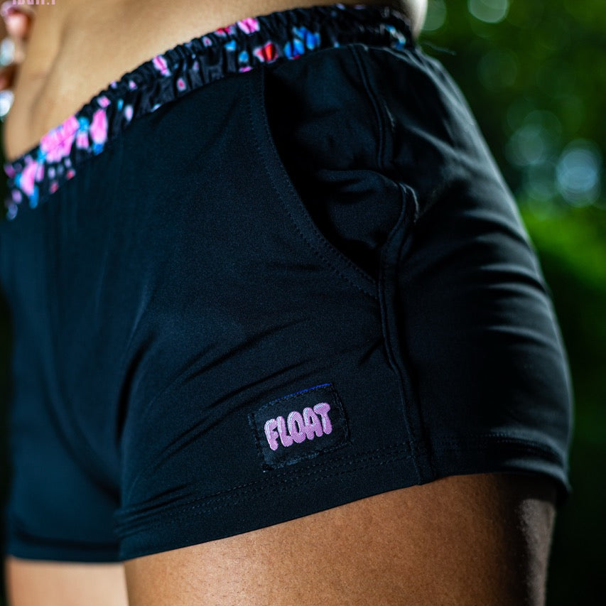 Close-up of a person wearing black athletic shorts with a colorful patterned waistband and a visible FLOAT logo patch on the side, photographed outdoors with blurred greenery in the background.