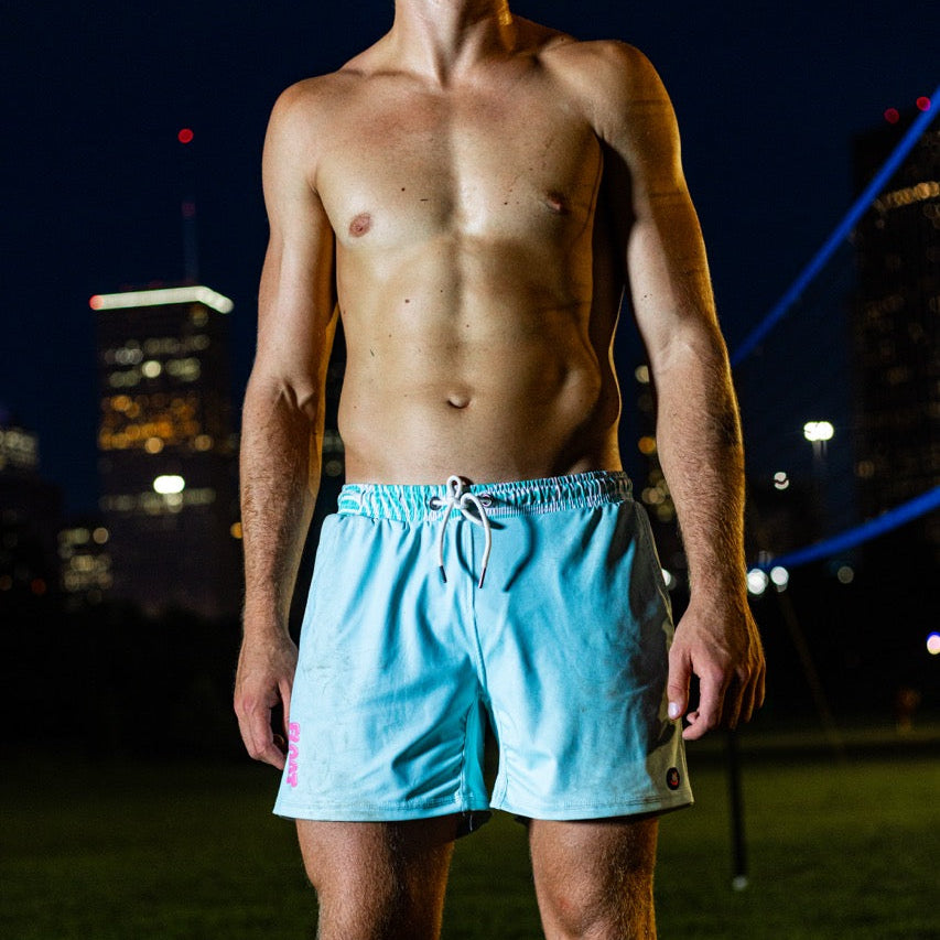 A shirtless man standing outdoors at night on grass, wearing light green athletic shorts with a white drawstring. City buildings and blue lights are visible in the background.