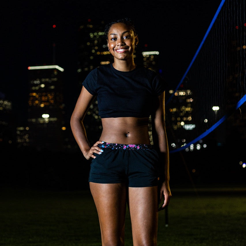 A woman stands outdoors at night wearing a black short-sleeve crop top and black athletic shorts with a colorful patterned waistband, photographed against a city skyline and bridge backdrop.