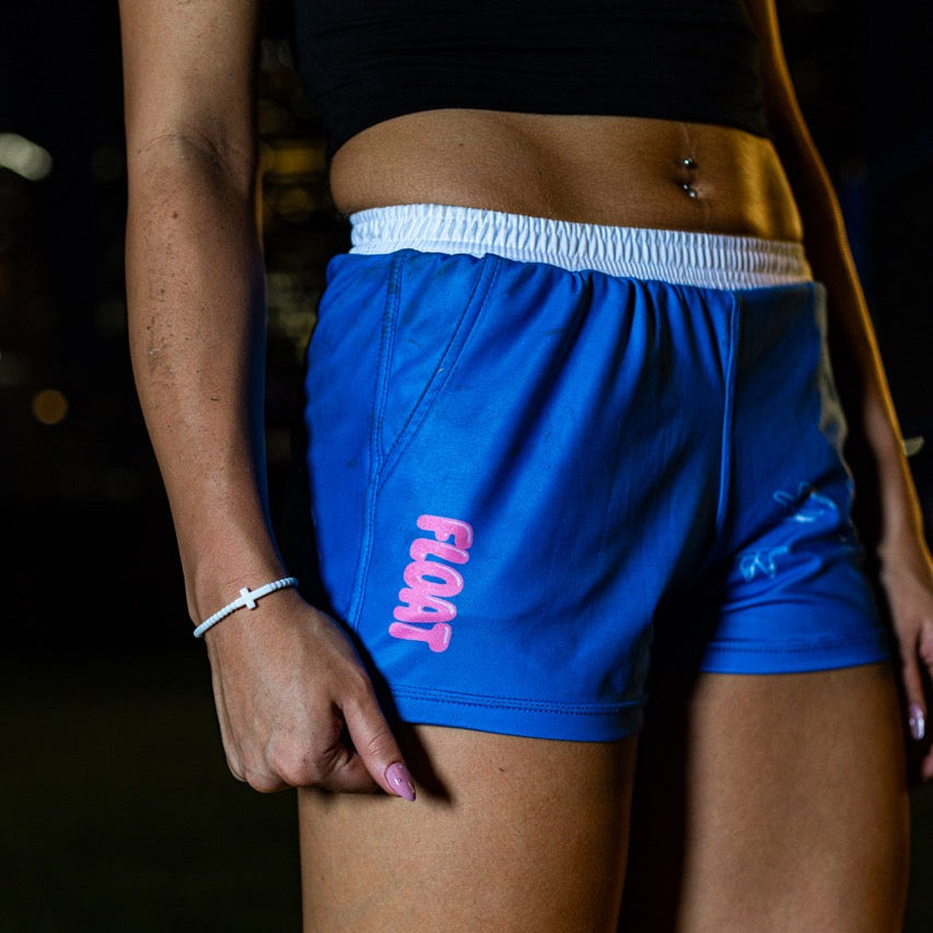 A close-up of a person wearing bright blue athletic shorts with a white elastic waistband and pink 'FLOAT' text on the left leg, paired with a black cropped top.
