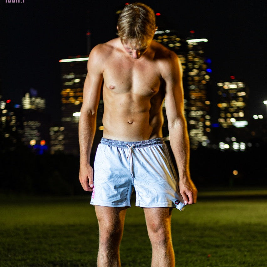 A shirtless man standing barefoot on grass at night with a blurred city skyline in the background, wearing light gray shorts.