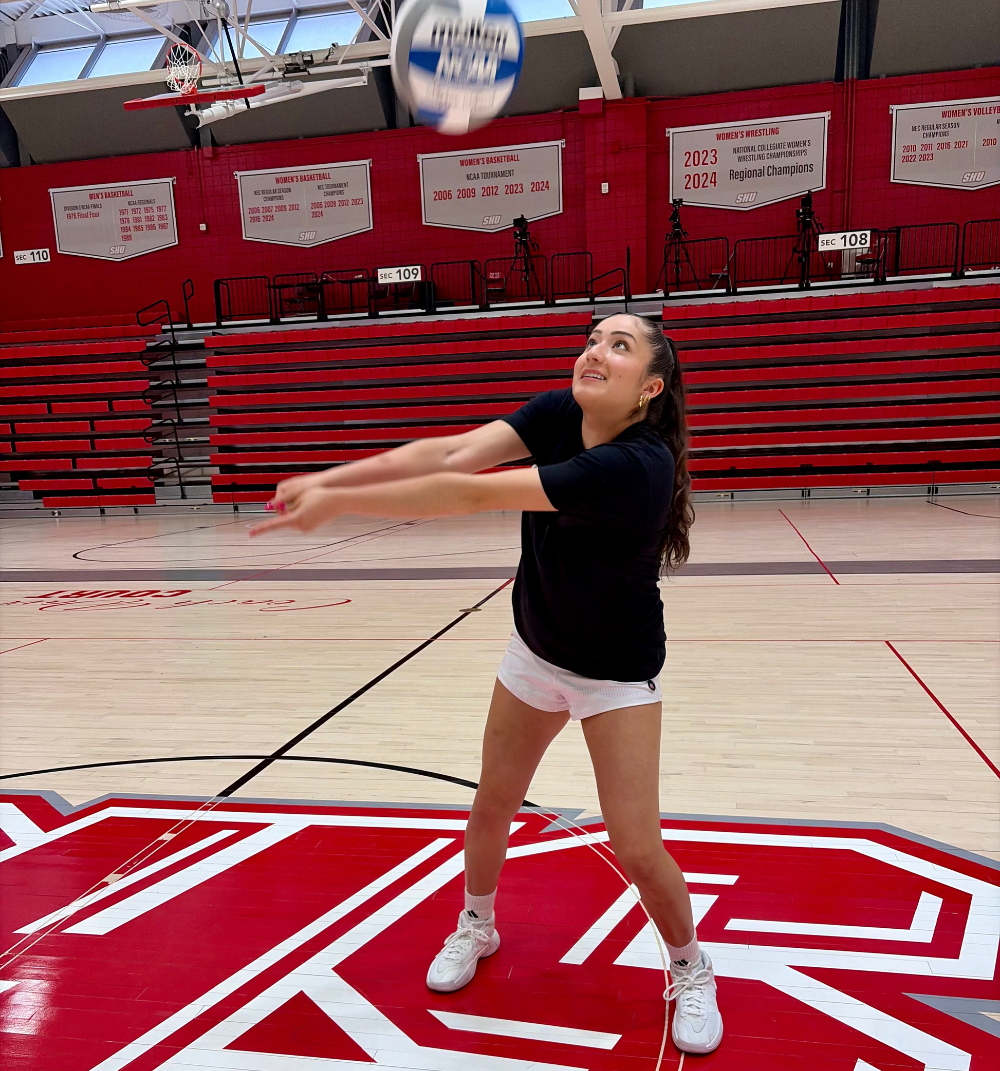 Person playing volleyball on a gymnasium floor with red bleachers and banners in the background.
