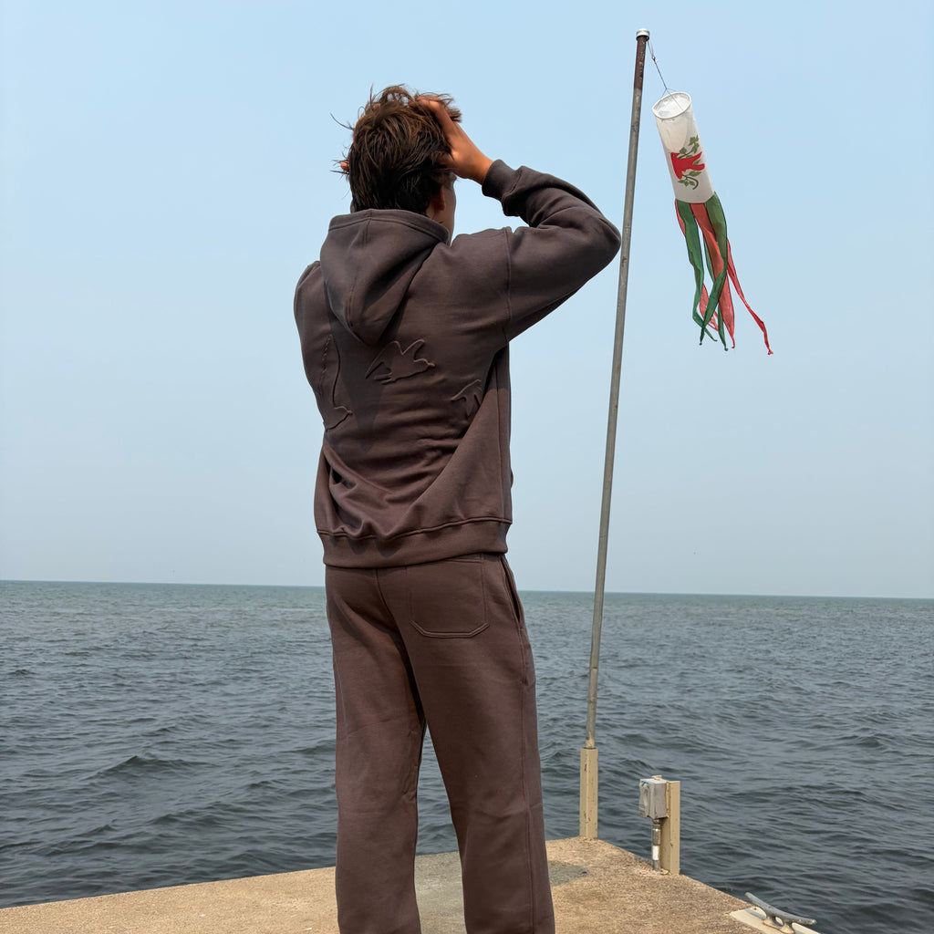 A person standing barefoot on a concrete pier with the ocean in the background, wearing a dark brown long-sleeve top and matching pants. A small white flag with red and green details is attached to a pole.
