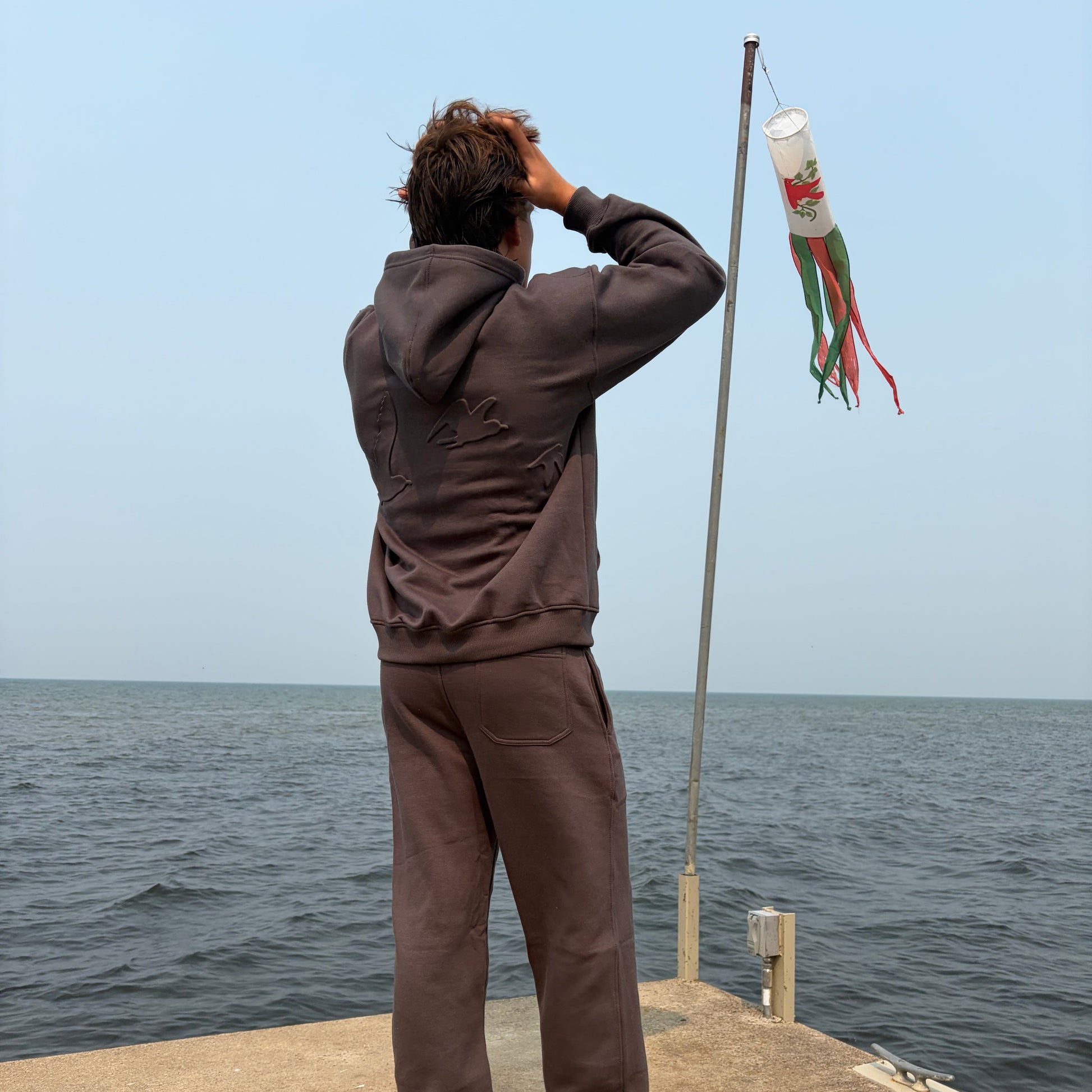 A person standing barefoot on a concrete pier with the ocean in the background, wearing a dark brown long-sleeve top and matching pants. A small white flag with red and green details is attached to a pole.