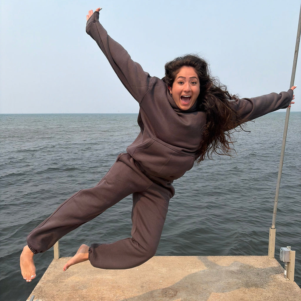 A woman in a matching gray tracksuit jumps dynamically on a concrete pier with the ocean in the background.