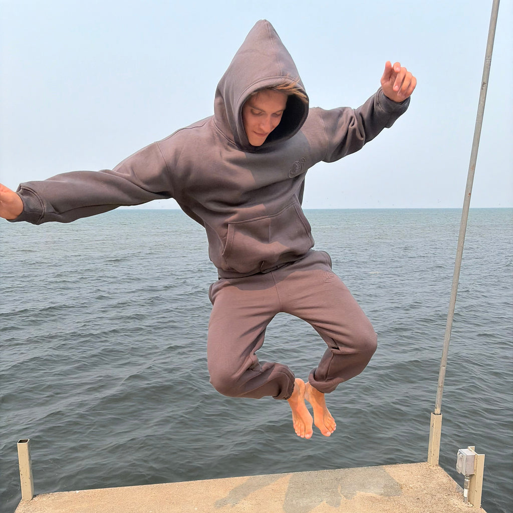 A person in a brownish-gray hoodie and matching sweatpants is captured mid-air while jumping off a concrete pier over water.