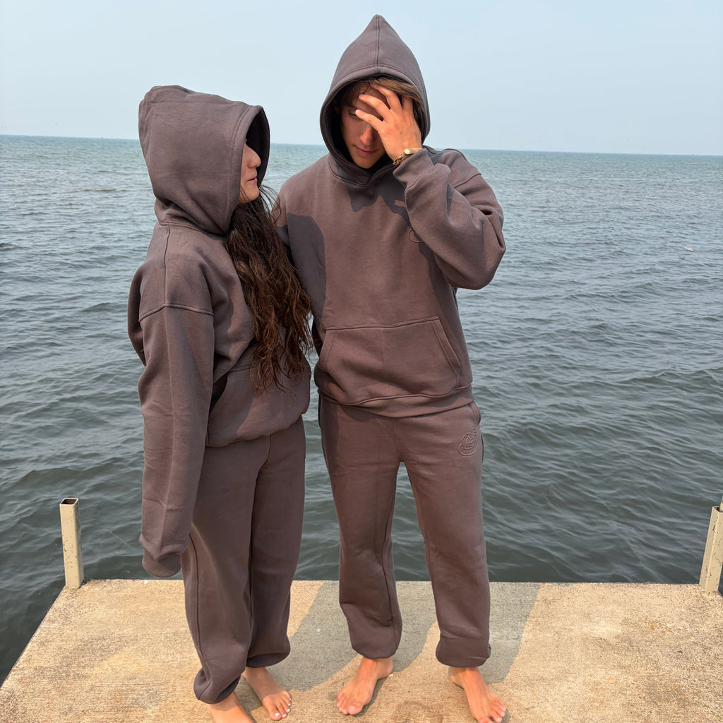 Two people standing barefoot on a concrete dock with the ocean in the background, wearing matching dark gray hooded sweatshirt and sweatpants sets.