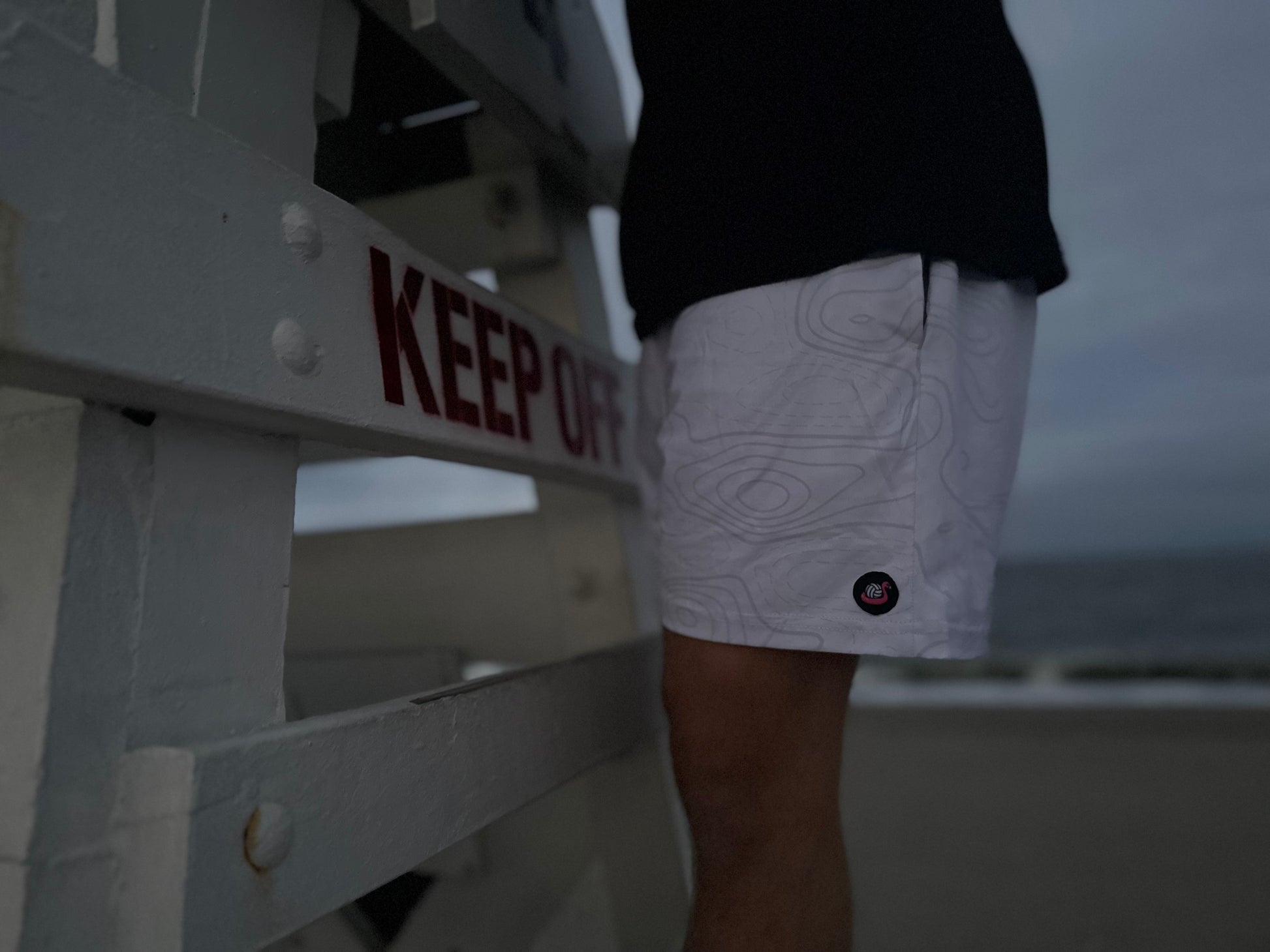 Person wearing white shorts with a pattern, standing next to a 'KEEP OFF' sign on a beach.