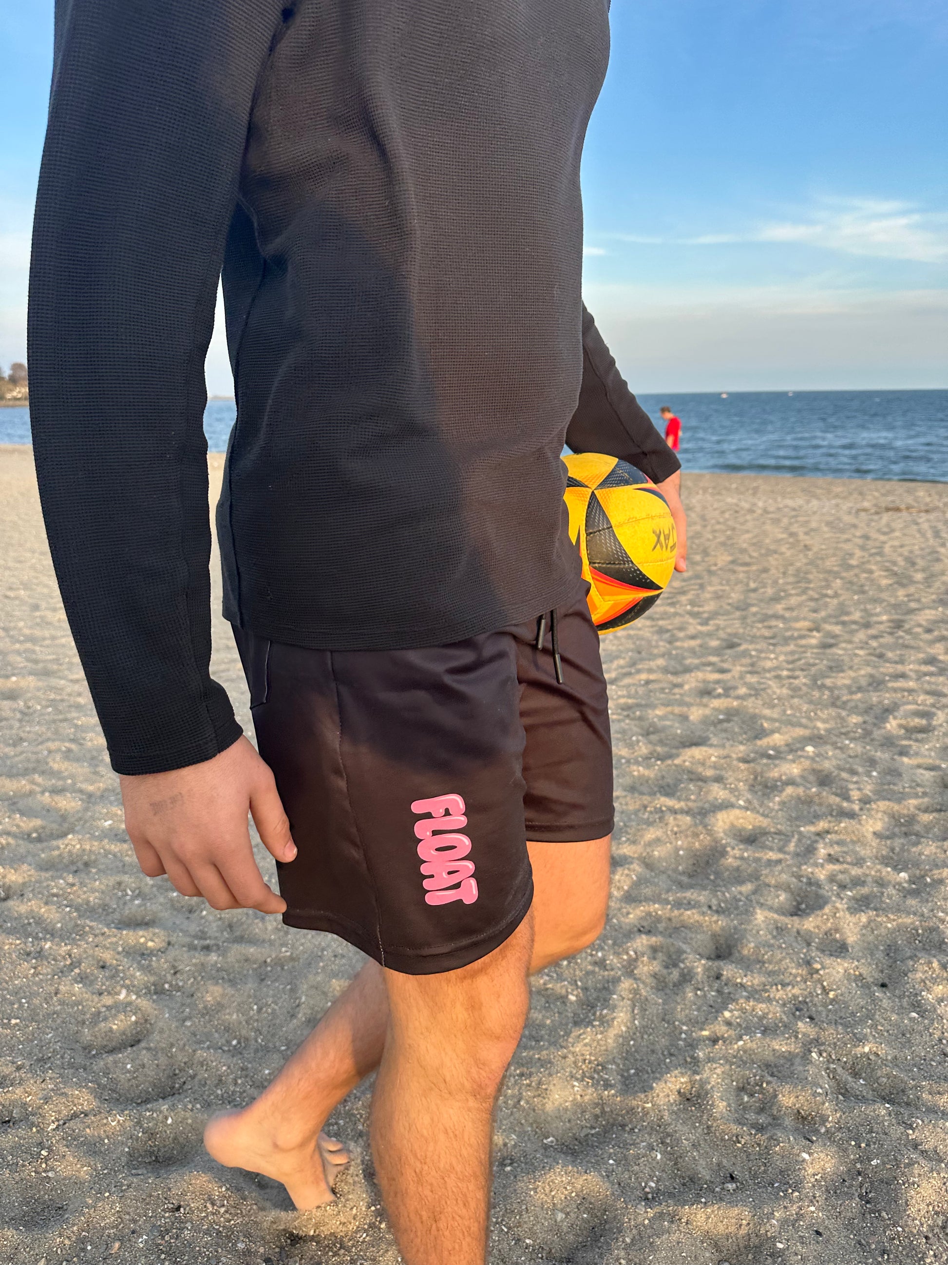 Person on a beach holding a sports ball, wearing black shorts with pink text.