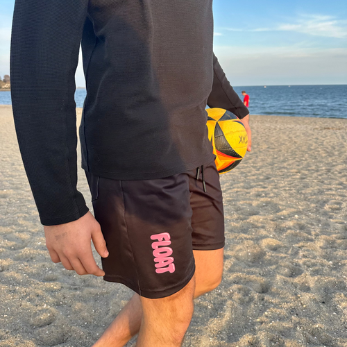 Person on a beach holding a sports ball, wearing black shorts with pink text.
