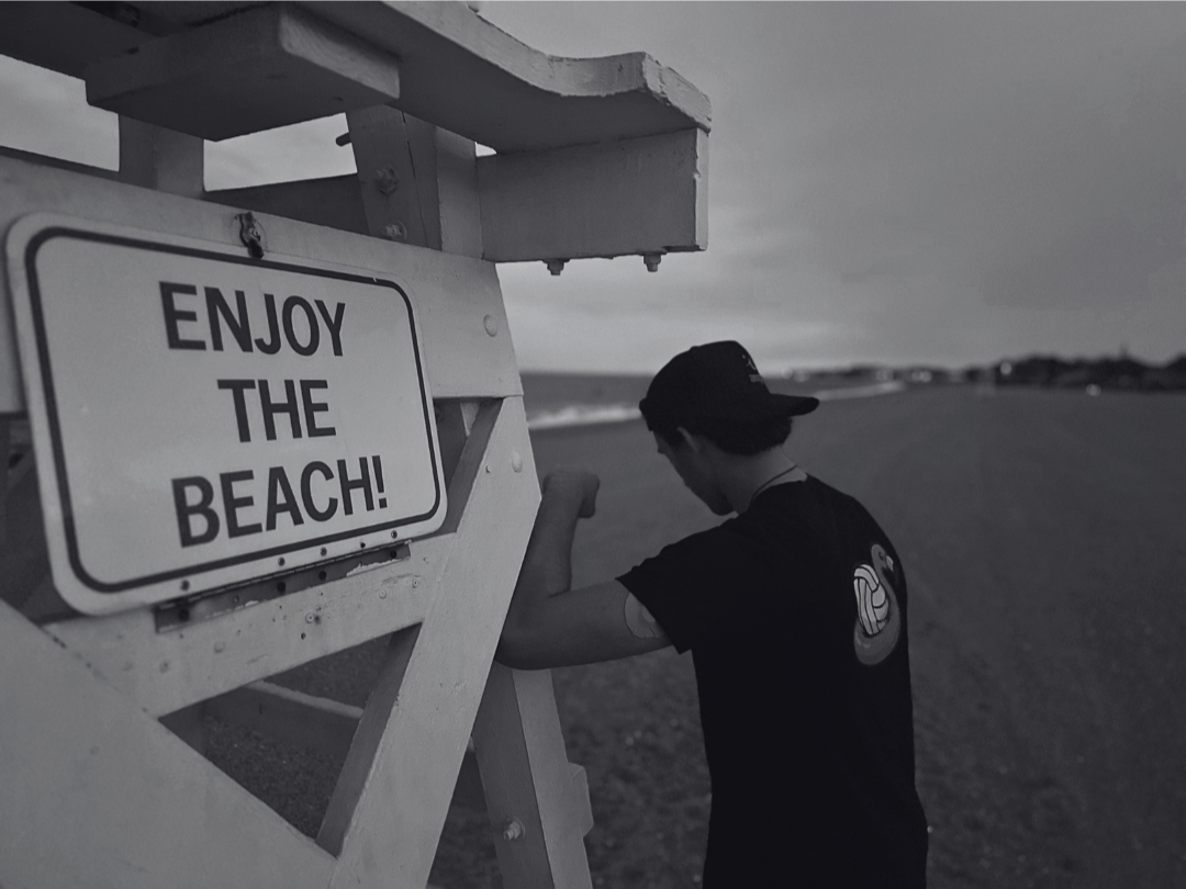Person standing next to a 'Enjoy the Beach!' sign on a beach.